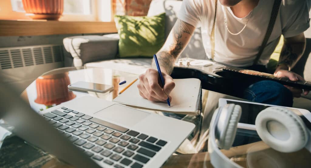 Um homem sentado em frente a um notebook enquanto consulta folhas de anotações sobre o curso online do MEC, com um lápis na mão 