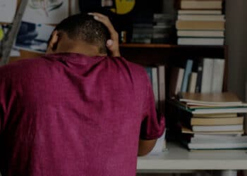 De costas um adolescente com a mão na cabeça enquanto estuda, demonstrando preocupação de não conseguir bolsa de estudo.