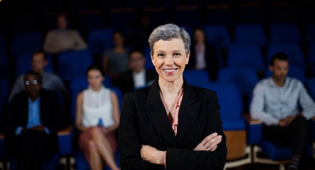 Uma mulher em cima de um placo sorri para a câmera com carisma. Atrás dela, em desfoque, vemos um auditório e várias pessoas acompanhando sua ação, mostrando a influência política do carisma.