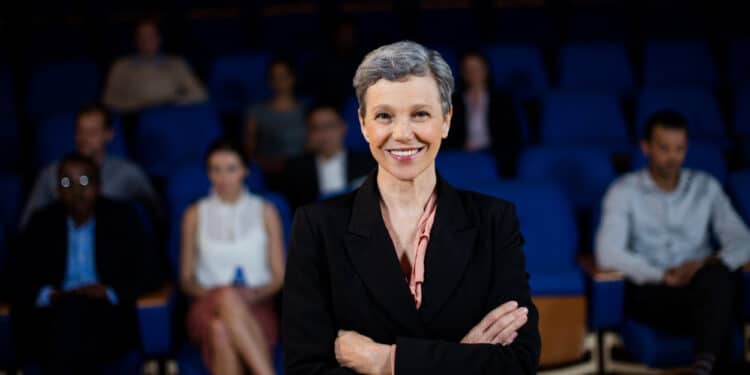 Uma mulher em cima de um placo sorri para a cГѓВўmera com carisma. AtrГѓВЎs dela, em desfoque, vemos um auditГѓВіrio e vГѓВЎrias pessoas acompanhando sua aГѓВ§ГѓВЈo, mostrando a influГѓВЄncia polГѓВ­tica do carisma.