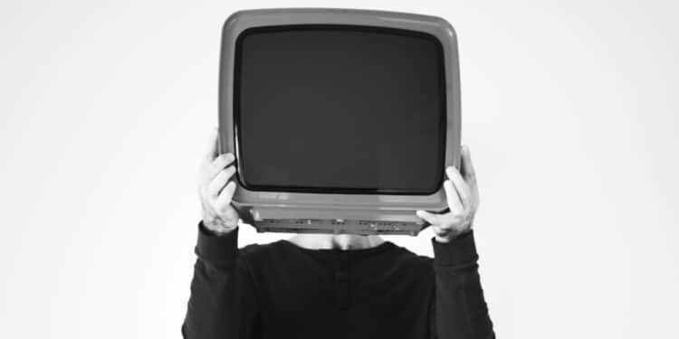 Um homem adulto estГѓВЎ de pГѓВ© e segura com as duas mГѓВЈos, na altura do rosto, um aparelho de televisГѓВЈo antigo na mГѓВ­dia.