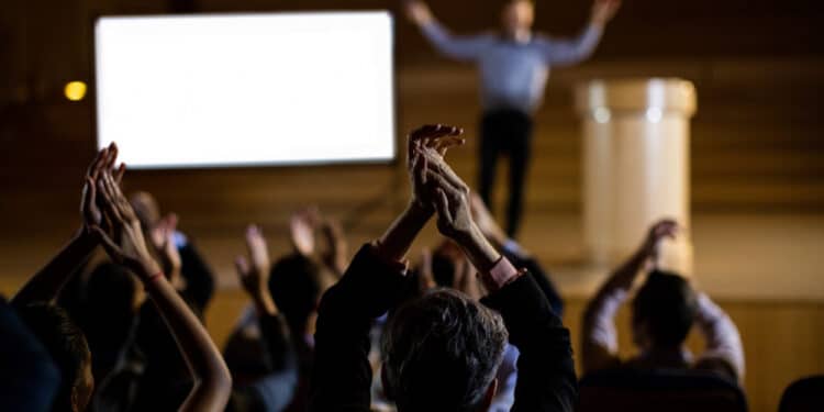 Em primeiro plano vemos diversas pessoas batendo palma viradas para um palco. Ao fundo, em desfoque, uma figura masculina com os braГѓВ§os para cima parece encerrar um discurso de poder.