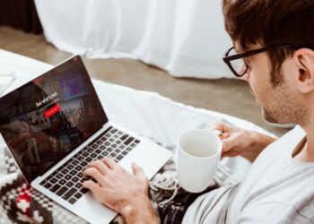 Homem segurando uma xícara de café e usando um laptop com a Netflix na tela na cama | Créditos: depositphotos.com / ViktoriaSapata