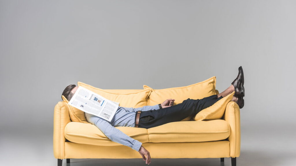 tired businessman sleeping on yellow sofa with newspaper on face, on grey