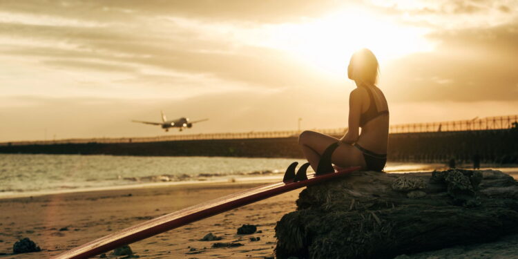 woman sitting on rock with surfboard on beach at sunset with airplane in sky