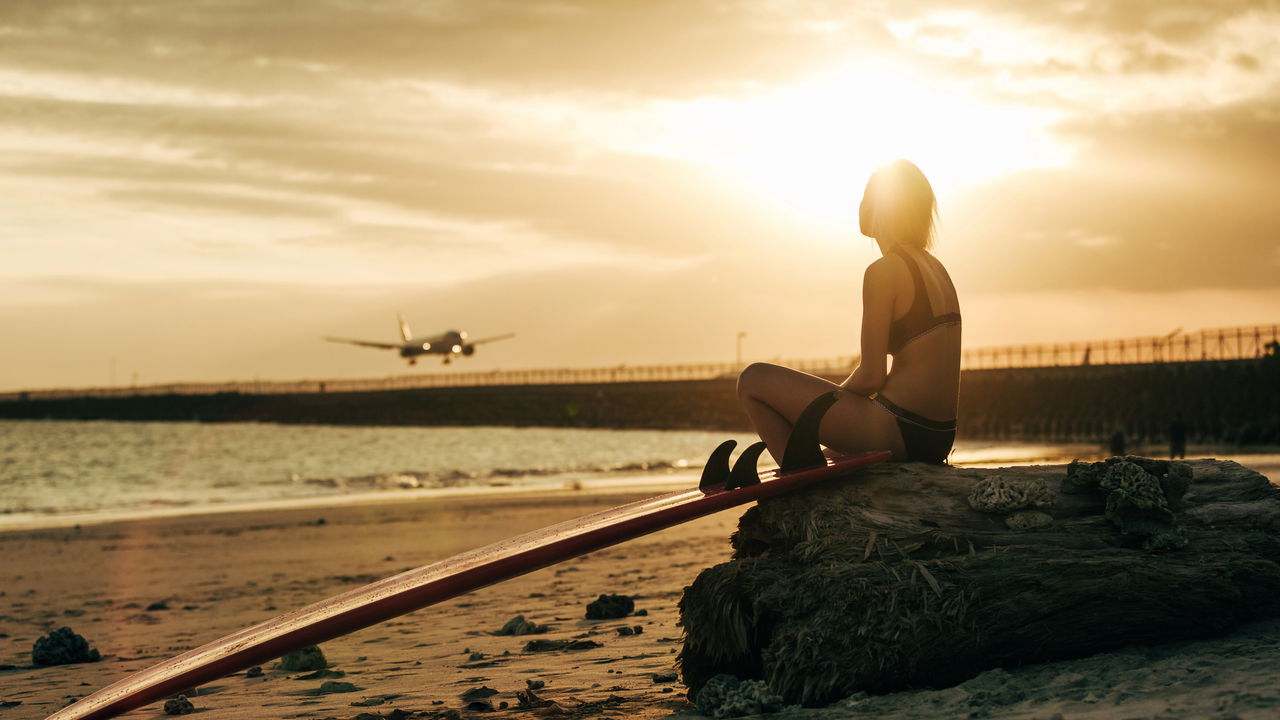 woman sitting on rock with surfboard on beach at sunset with airplane in sky