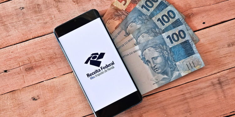 Cédulas de real próximas a celular com logo da Receita Federal - Créditos: depositphotos.com / Alisonnunes