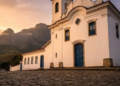 A histórica Matriz de Santo Antônio sob a luz dourada, com as imponentes montanhas da Serra ao fundo.