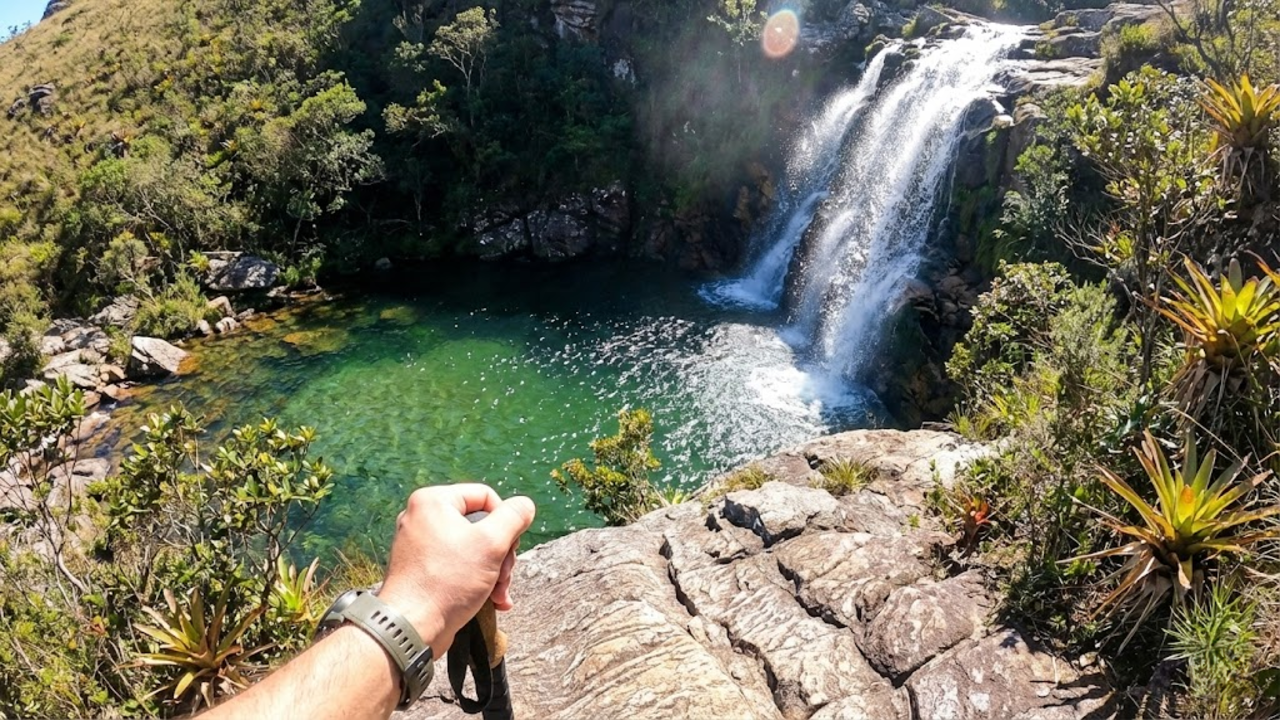 Visão em primeira pessoa de um trilheiro contemplando a queda d'água cristalina da Cachoeira de Itatiaia.
