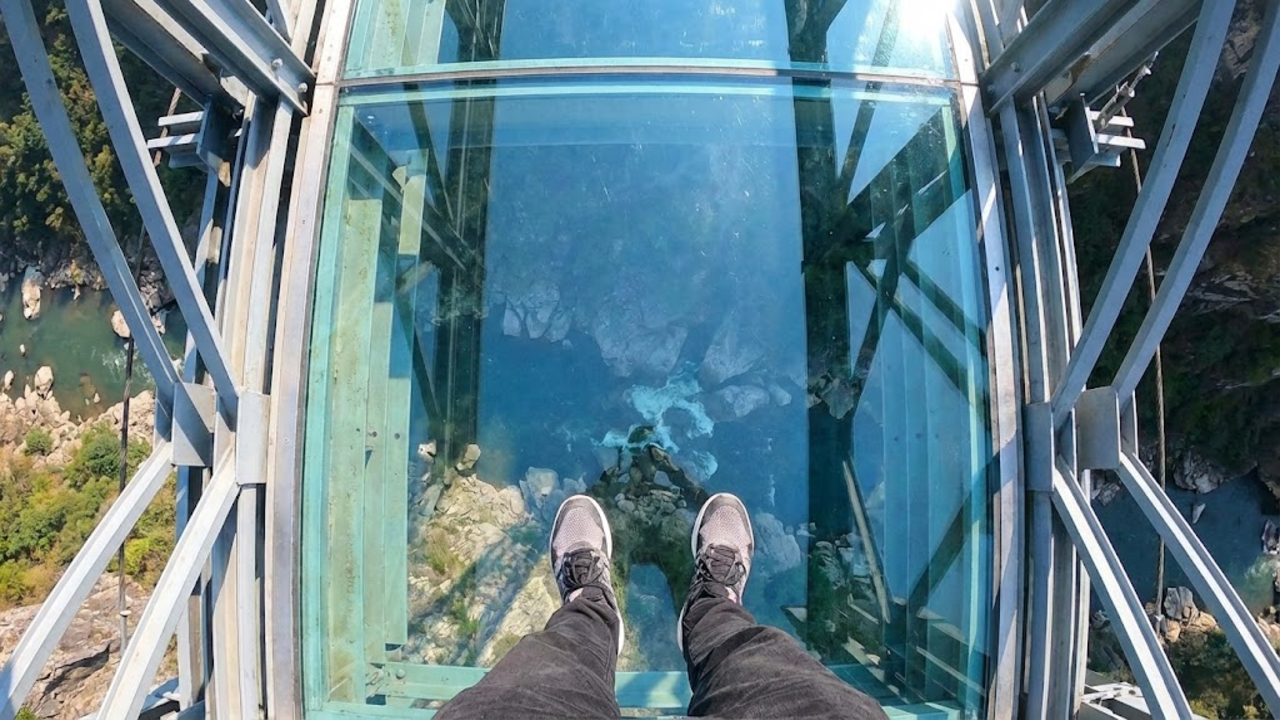 Visão em primeira pessoa olhando para baixo através do piso de vidro da ponte, revelando o abismo de 625 metros.