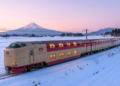 O trem Sunrise Express atravessando uma paisagem de neve ao amanhecer, com o Monte Fuji ao fundo e janelas iluminadas.