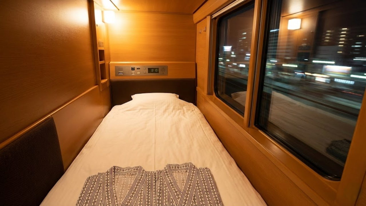 Ponto de vista de dentro da cabine 'Solo', mostrando a cama, o yukata dobrado e a janela com luzes da cidade à noite