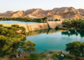 Uma vista panorâmica e dourada da barragem de pedra criando um lago azul cercado de verde no meio do deserto rochoso