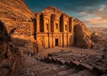 O Mosteiro Ad Deir é o maior e mais imponente monumento da cidade histórica de Petra, na Jordânia. Esculpido diretamente na rocha sólida no século I, o templo possui 47 metros de largura e é um testemunho da genialidade nabateia. Onde fica o Mosteiro e como é o acesso pela montanha? Diferente do famoso Tesouro (Al-Khazneh), que fica logo na entrada, o Mosteiro está localizado no alto das montanhas de Petra. Para alcançá-lo, o visitante deve subir uma escadaria de aproximadamente 800 degraus esculpidos na pedra. A subida é cansativa, mas recompensada por vistas panorâmicas do vale de Araba. É um trajeto que exige preparo físico e calçados adequados, levando o turista para uma área menos congestionada e mais selvagem do sítio arqueológico jordaniano. Fachada monumental esculpida em rocha sólida nas montanhas de Petra no território jordaniano - Créditos: depositphotos.com / DI-blr Qual a diferença entre o Tesouro e o Mosteiro de Petra? Embora ambos apresentem fachadas monumentais, o Mosteiro é significativamente maior que o Tesouro e possui um design mais austero. Suas colunas e nichos são mais simples, focando na escala massiva e na perfeição das proporções arquitetônicas. Para que você compreenda as dimensões e a escala destes dois ícones, preparamos uma comparação técnica baseada nas medidas oficiais dos monumentos: Monumento Largura (Metros) Altura (Metros) Destaque Visual Tesouro (Al-Khazneh) 25 39 Detalhes ornamentais finos Mosteiro (Ad Deir) 47 48 Escala colossal e isolamento Leia também: Rodovia na Áustria ganha fama por sua inclinação de 12%, pois o trajeto a 1.893 metros de altitude une vales alpinos através de curvas fechadas e íngremes Por que o monumento recebeu o nome de Mosteiro? Apesar do nome "Mosteiro", a estrutura foi originalmente construída como um templo religioso ou túmulo real nabateu. O nome atual deriva de cruzes esculpidas nas paredes internas durante o período bizantino, quando o local foi utilizado por monges cristãos. O interior é composto por uma câmara simples e austera, contrastando com a grandiosidade da fachada externa. Segundo o portal oficial Visit Jordan, o local era palco de festivais religiosos importantes para o povo nabateu. Para se aprofundar na história de uma das sete maravilhas do mundo moderno, selecionamos o conteúdo do canal Foca na História, que conta com mais de 1 milhão de inscritos. No vídeo a seguir, o canal detalha a engenharia dos nabateus e as impressionantes estruturas esculpidas na pedra, como o famoso Monastério de Petra: Quais os indicadores oficiais do sítio arqueológico de Petra? Entender os dados de Petra ajuda a dimensionar a importância deste Patrimônio Mundial da UNESCO. A cidade rosa-vermelha é o motor do turismo na Jordânia, recebendo milhões de pessoas que buscam a história das civilizações antigas do deserto. Segundo os dados de turismo do Governo da Jordânia, os indicadores locais são: Extensão do Parque: Aproximadamente 264 quilômetros quadrados. Patrimônio: Considerada uma das Sete Maravilhas do Mundo Moderno. Visitantes: Média de 1 milhão de pessoas por ano. Idade: Fundada por volta do século IV a.C. pelos Nabateus. Qual o melhor horário para fotografar o Mosteiro Ad Deir? O final da tarde é o momento ideal para visitar o Mosteiro, quando a luz do sol ilumina diretamente a fachada dourada. Nesse horário, a maioria das excursões já iniciou o retorno, permitindo uma experiência mais silenciosa e contemplativa. No topo, existem pequenos cafés beduínos onde o turista pode descansar e apreciar a magnitude da obra. O Mosteiro Ad Deir permanece como o maior símbolo da audácia de um povo que transformou montanhas brutas em monumentos de beleza eterna na Jordânia.