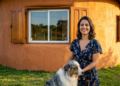 Arquiteta sorrindo com seu cachorro em frente à sua bela casa rústica de paredes curvas de barro sob a luz do pôr do sol.