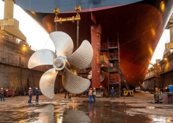 Hélice gigante de navio cargueiro é instalada em estaleiro com guindastes e casco ao fundo