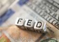 Cubos metálicos formam a palavra “FED” sobre uma nota de dólar americano, em referência ao Federal Reserve e à política monetária dos Estados Unidos.
