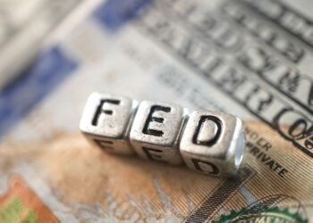 Cubos metálicos formam a palavra “FED” sobre uma nota de dólar americano, em referência ao Federal Reserve e à política monetária dos Estados Unidos.