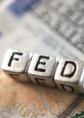 Cubos metálicos formam a palavra “FED” sobre uma nota de dólar americano, em referência ao Federal Reserve e à política monetária dos Estados Unidos.