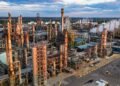 Vista aérea de uma refinaria de petróleo, com torres de processamento, tubulações e estruturas industriais.