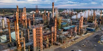 Vista aérea de uma refinaria de petróleo, com torres de processamento, tubulações e estruturas industriais.