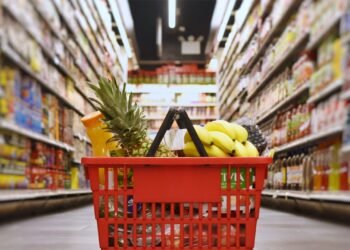 Cesta de compras com frutas e outros produtos em corredor de supermercado.
