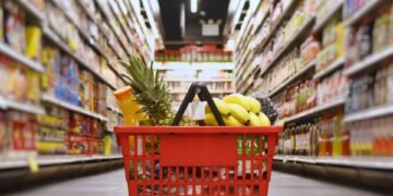 Cesta de compras com frutas e outros produtos em corredor de supermercado.